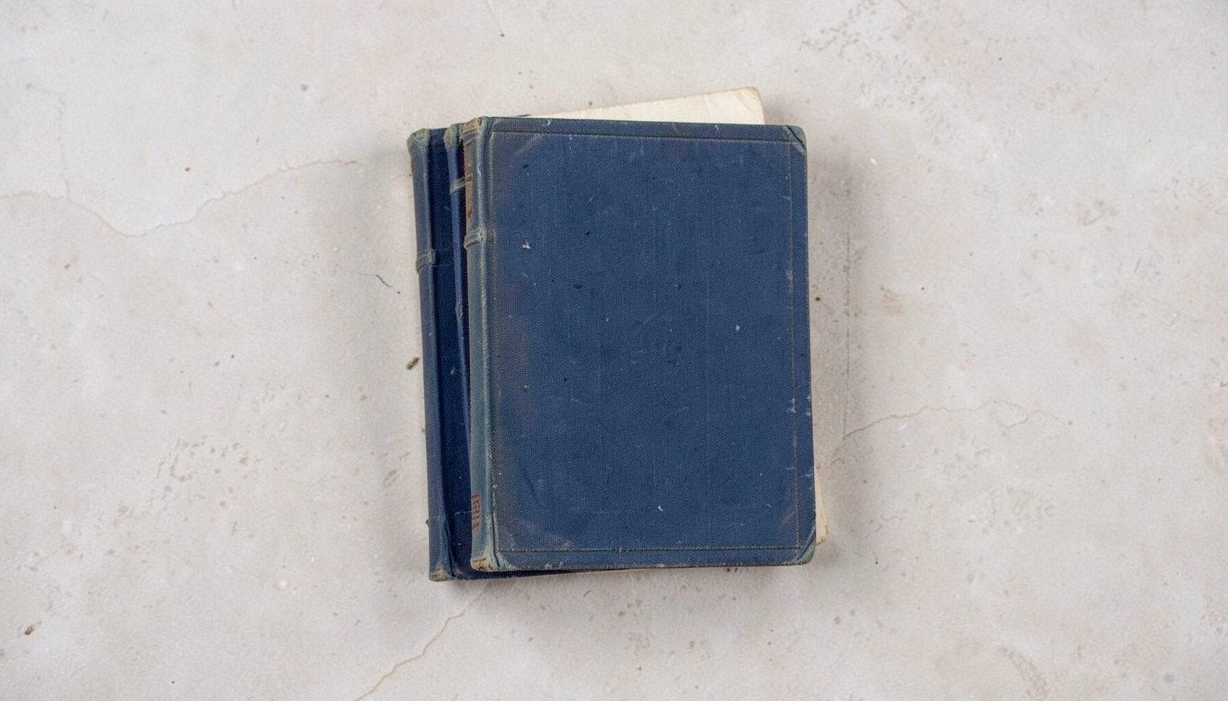 Overhead flat-lay photograph of three stacked archival books with worn cloth bindings in muted blues and creams, resting on a pale stone surface, conveying a quiet, documentary mood.