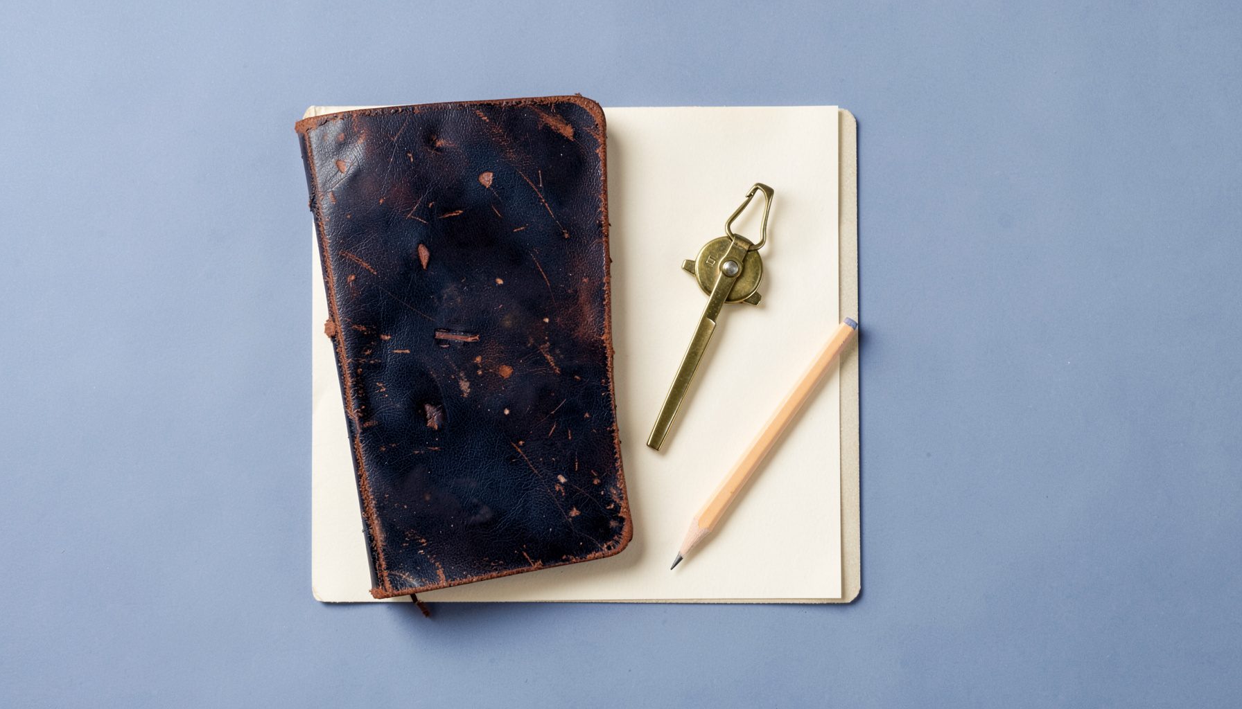 Overhead flat-lay photograph of a worn leather-bound notebook, a brass compass, and a pencil on cream paper, viewed from above with soft shadows.