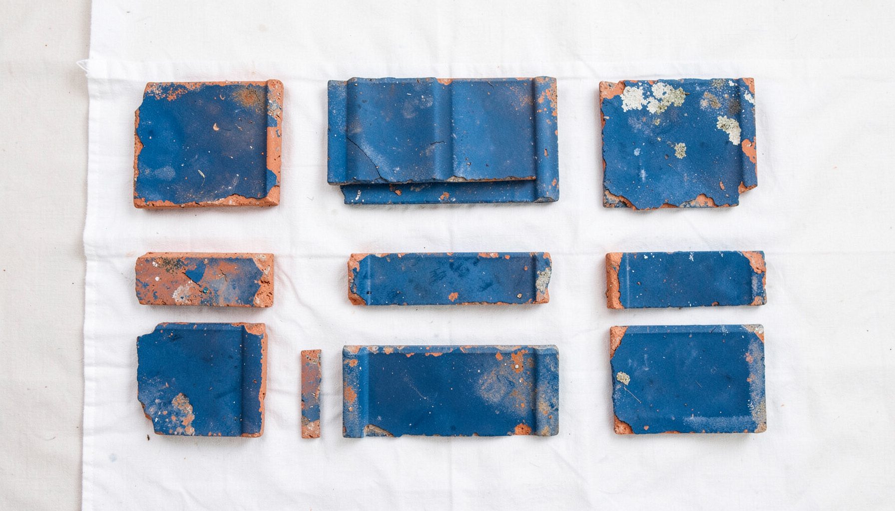 Overhead flat-lay photograph of weathered terracotta roof fragments and faded blue tiles arranged in a loose grid on white linen.