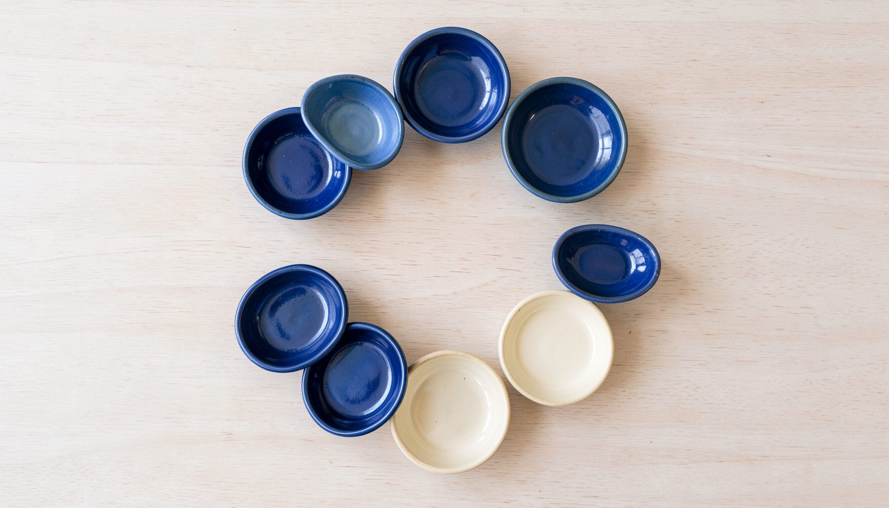 Overhead flat-lay photograph of a collection of small ceramic dishes in cobalt blue and warm ivory arranged in a circle on pale wood.
