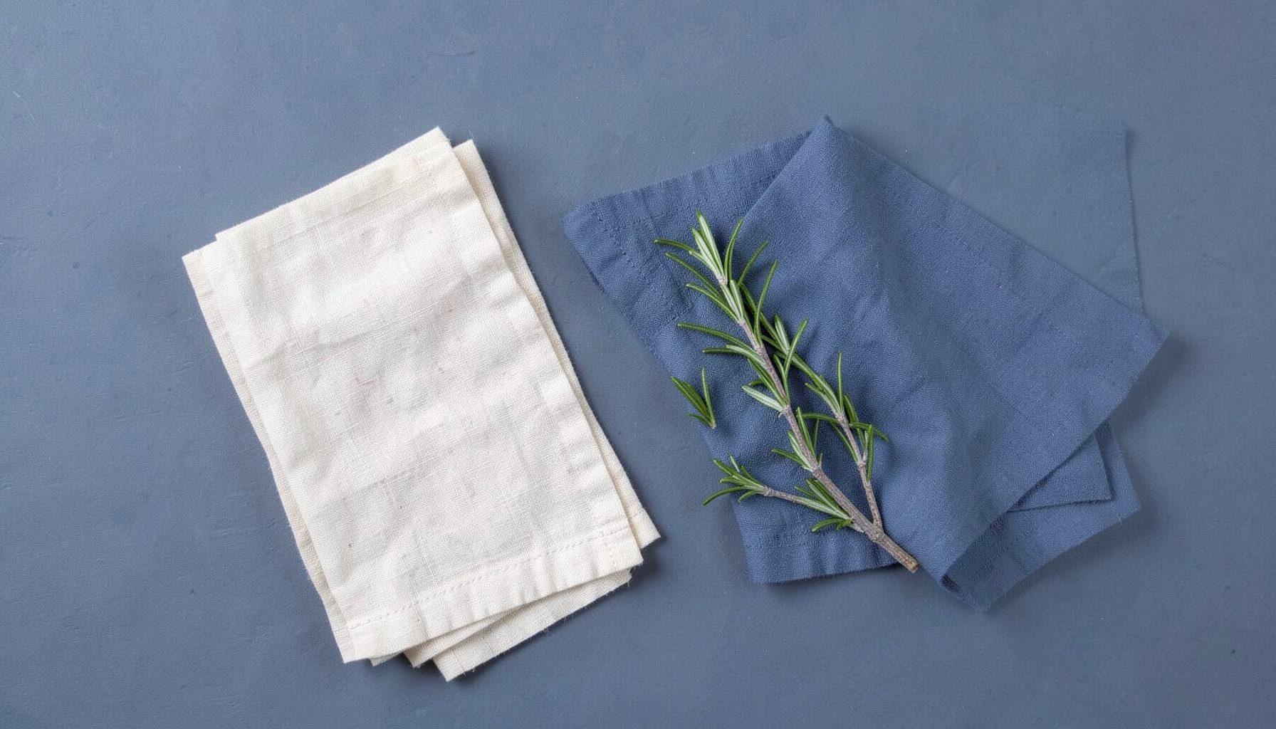 Overhead flat-lay photograph of neatly folded cream linen napkins beside a sprig of dried rosemary, viewed from above on a slate-grey background.