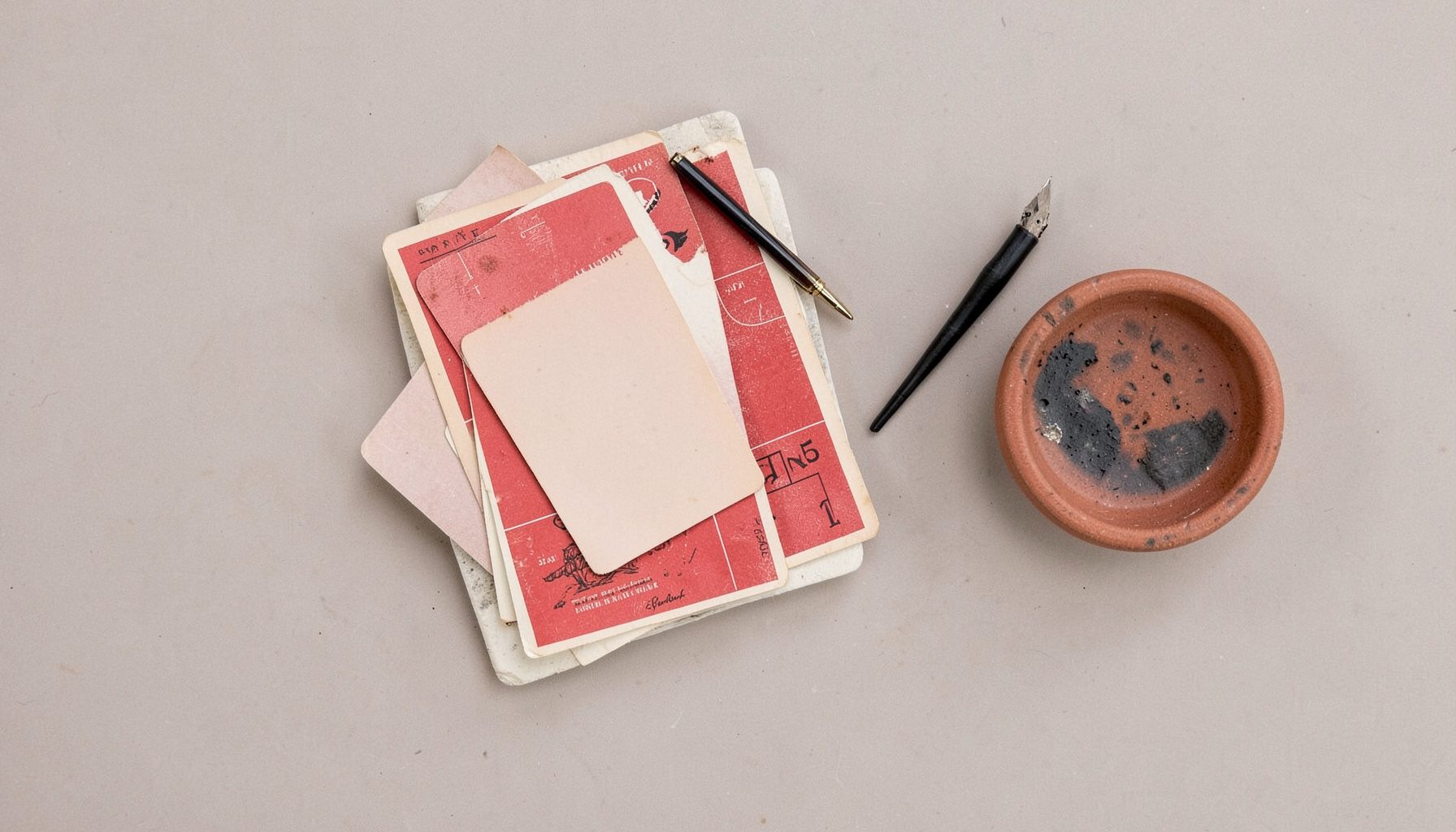 Overhead flat-lay photograph of a stone counter with a stack of vintage postcards, a fountain pen, and a small ceramic dish, in dusty terracotta, ivory, and charcoal.