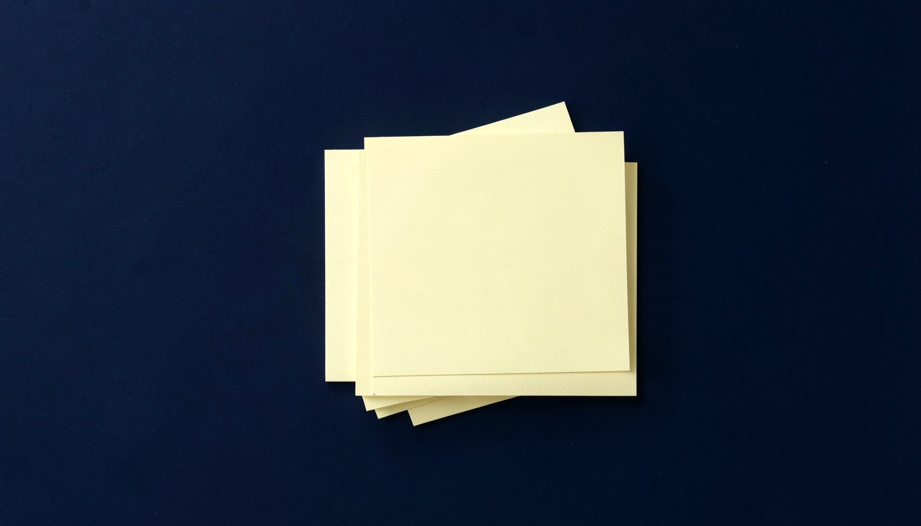 Overhead flat-lay photograph of pale yellow stationery cards stacked neatly on a dark navy background, soft daylight from one side.
