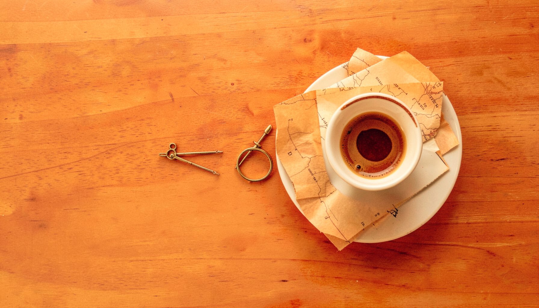 Overhead flat-lay photograph of a polished wooden table with a small brass compass, a folded paper map, and a cup of espresso on a saucer, in warm amber and cream tones.