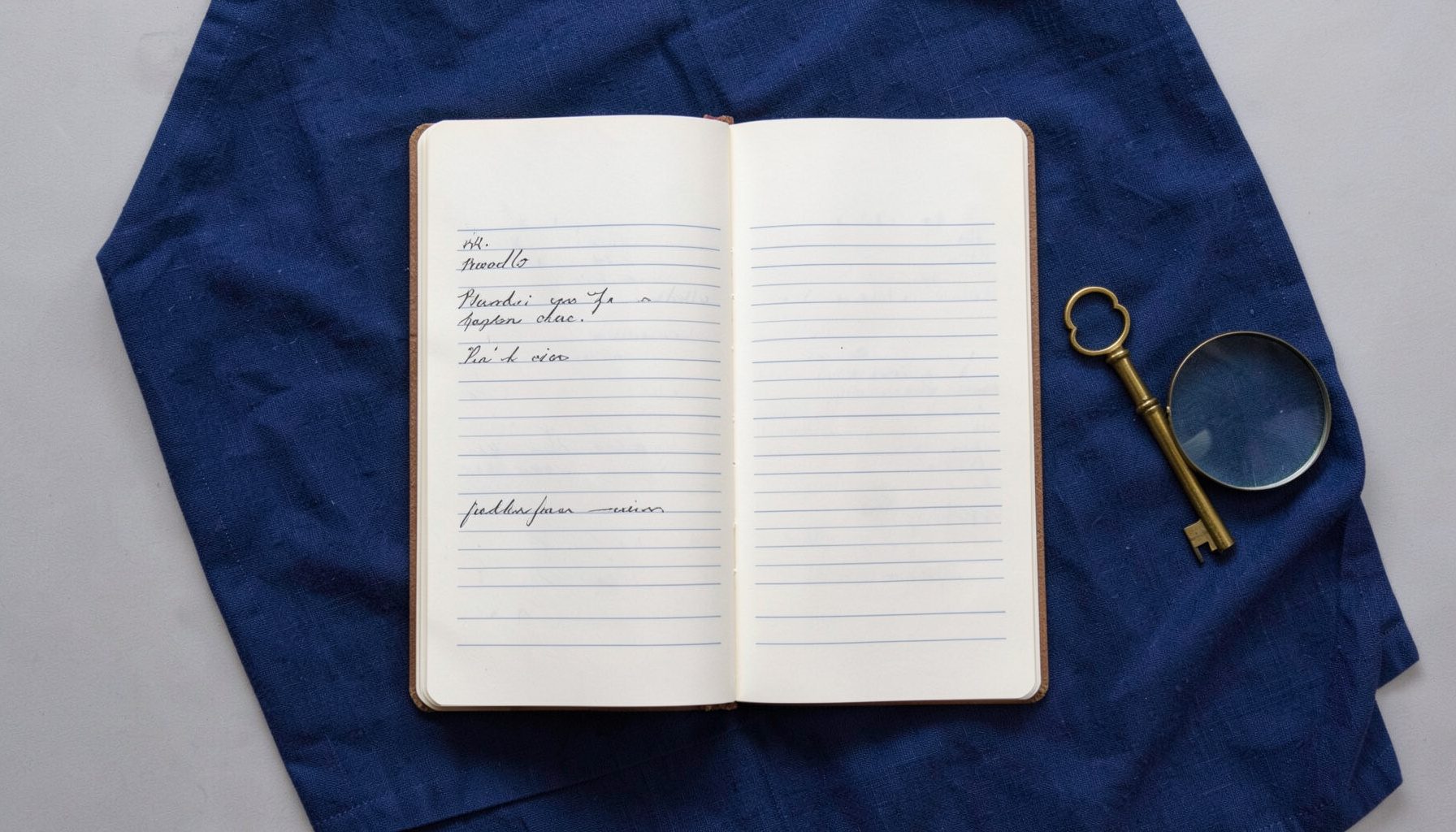 Overhead flat-lay photograph of a single open ledger with handwritten lines, a brass key, and a small magnifying glass on a navy cloth.