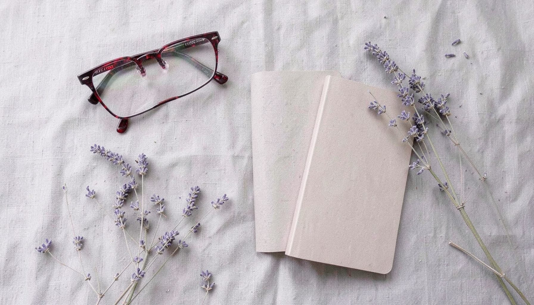 Overhead flat-lay photograph of a linen surface with a pair of reading glasses, a slim notebook, and dried lavender sprigs in soft grey, beige, and muted violet.